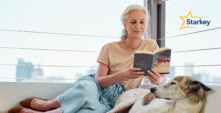 Image of woman reading a book beside her dog and a window